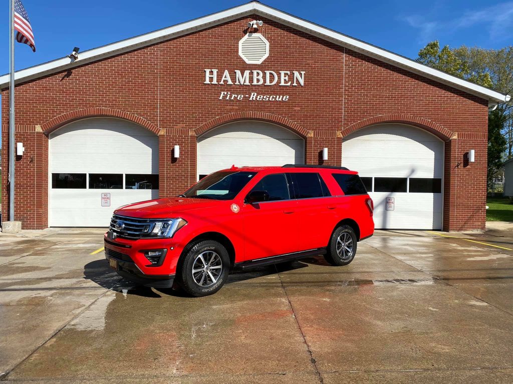 A bright red SUV parked in front of a brick building labeled "Hambden Fire-Rescue." The building has three white garage doors and an American flag on a pole to the left. The ground is wet, reflecting the sunny blue sky.