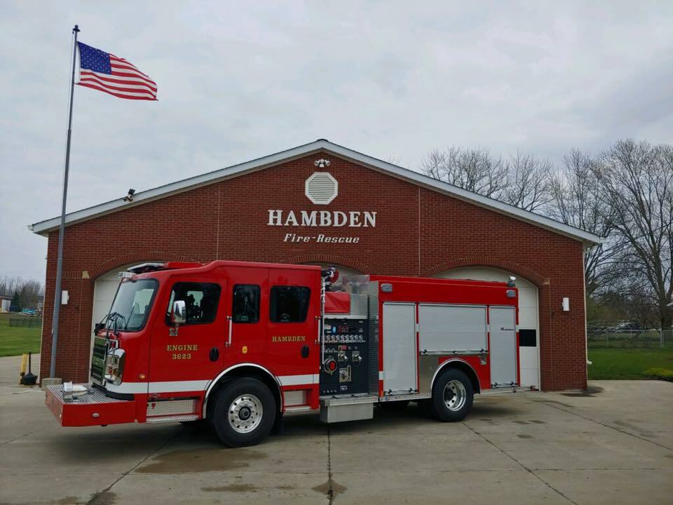 A red fire engine is parked in front of a brick fire station building labeled "HAMBDEN FIRE-RESCUE." An American flag is flying on a pole nearby. The sky is overcast, and trees are visible in the background.