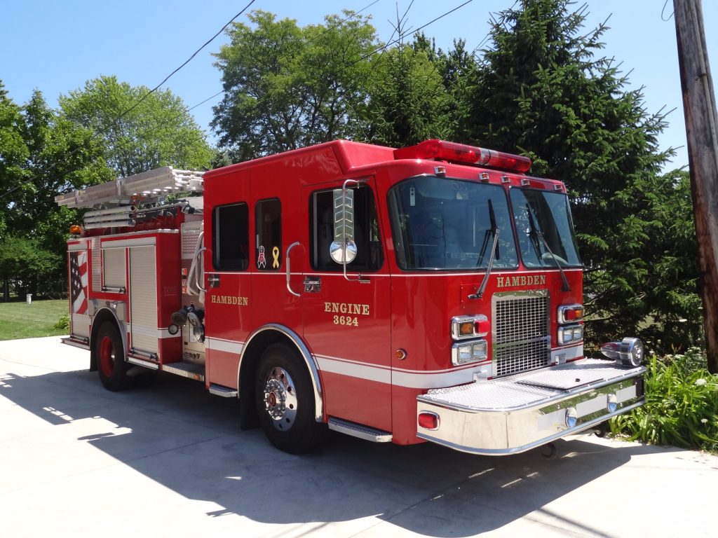 A bright red fire truck with "HAMBDEN" and "ENGINE 3624" written on it is parked on a paved driveway. It is equipped with ladders, hoses, and emergency lights. Trees and a utility pole are visible in the background under a clear sky.