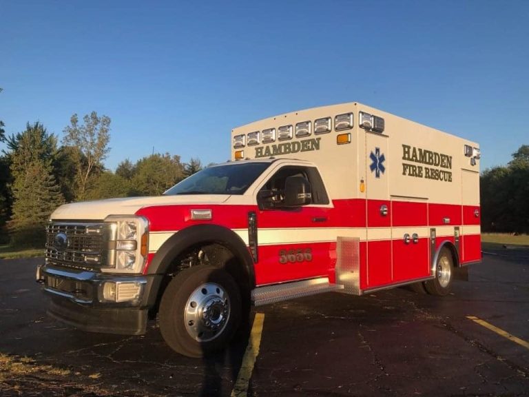 A red and white ambulance labeled "Hambden Fire Rescue" is parked on a paved surface. The vehicle is under a clear blue sky, surrounded by trees, and is illuminated by warm sunlight.