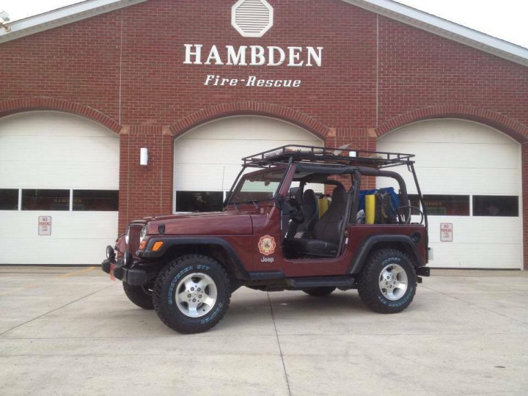 A maroon Jeep with a roll cage and off-road lights is parked in front of the Hambden Fire-Rescue station. The building features three garage doors and a brick facade.