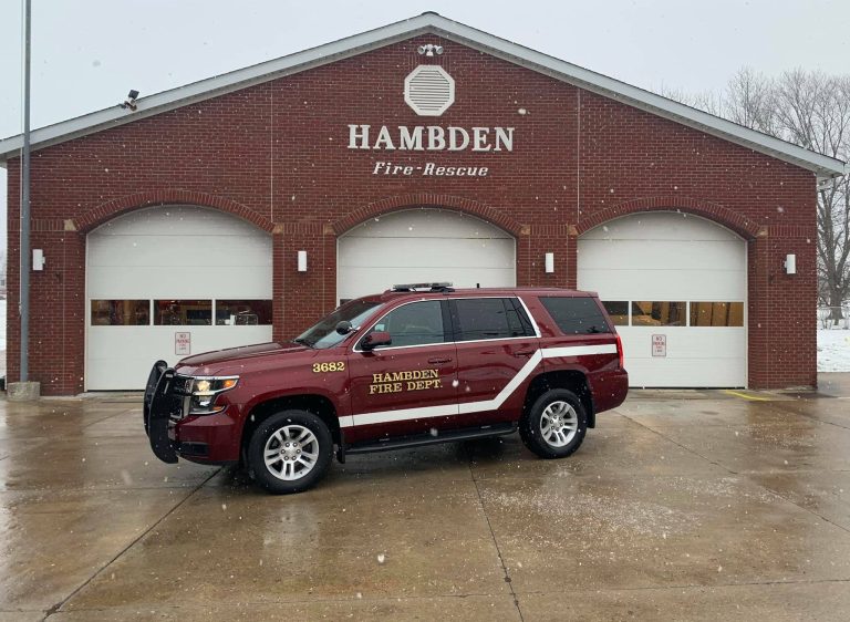 A red fire department SUV is parked in front of the brick building of the Hambden Fire Rescue. Snow is falling lightly, and the garage doors behind the vehicle are closed.