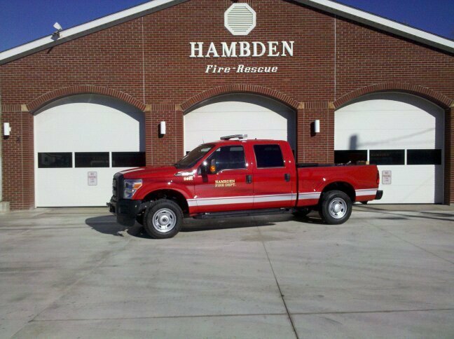 A red Hamden Fire Department truck is parked in front of a brick fire station with three large garage doors. The station has "HAMBDEN Fire Rescue" written above the doors.