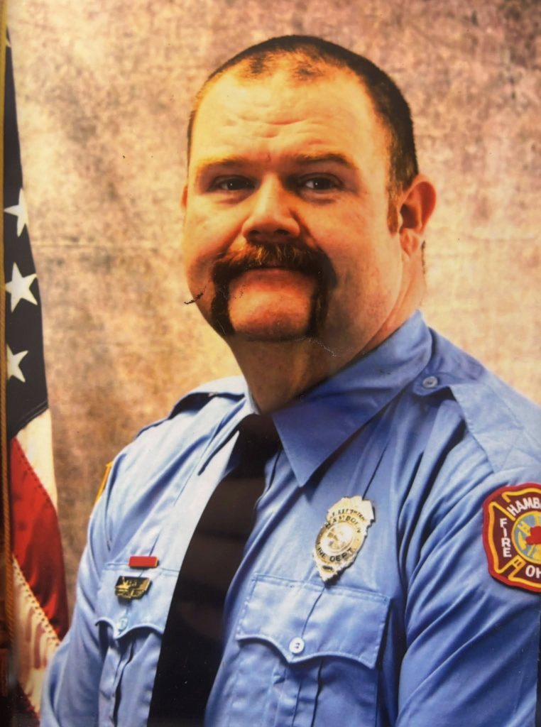 A firefighter with a handlebar mustache wears a blue uniform and black tie, with badges on his chest and a patch on his sleeve. He poses in front of a U.S. flag and mottled brown background.