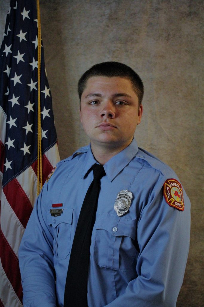 A young person in a light blue uniform stands in front of an American flag. They are wearing a tie and a badge on their shirt, which also has a patch on the shoulder. The background is a neutral, textured panel.