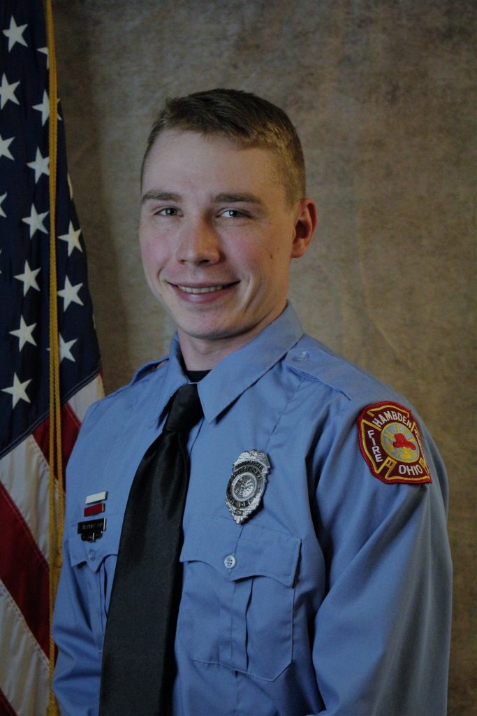 A person in a light blue uniform with a black tie and a badge stands smiling in front of an American flag. The uniform has a red and yellow patch on the left shoulder.