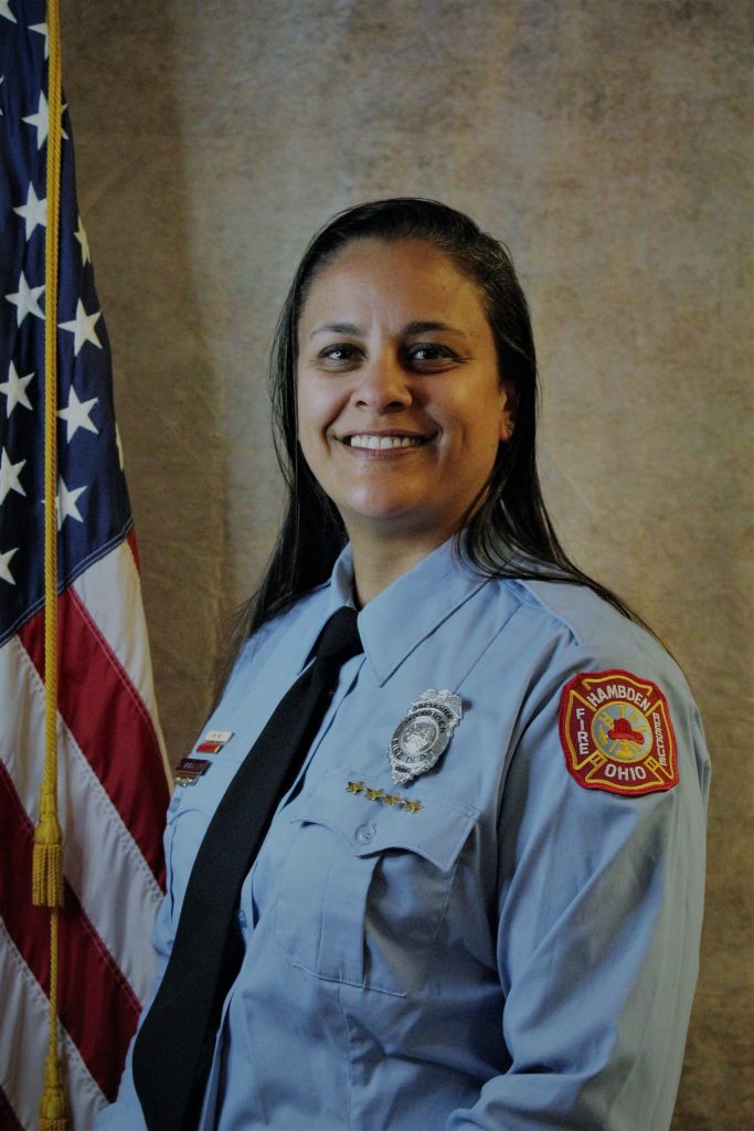 A person in a light blue uniform with a badge and patches stands smiling in front of the American flag. The background is a neutral color.
