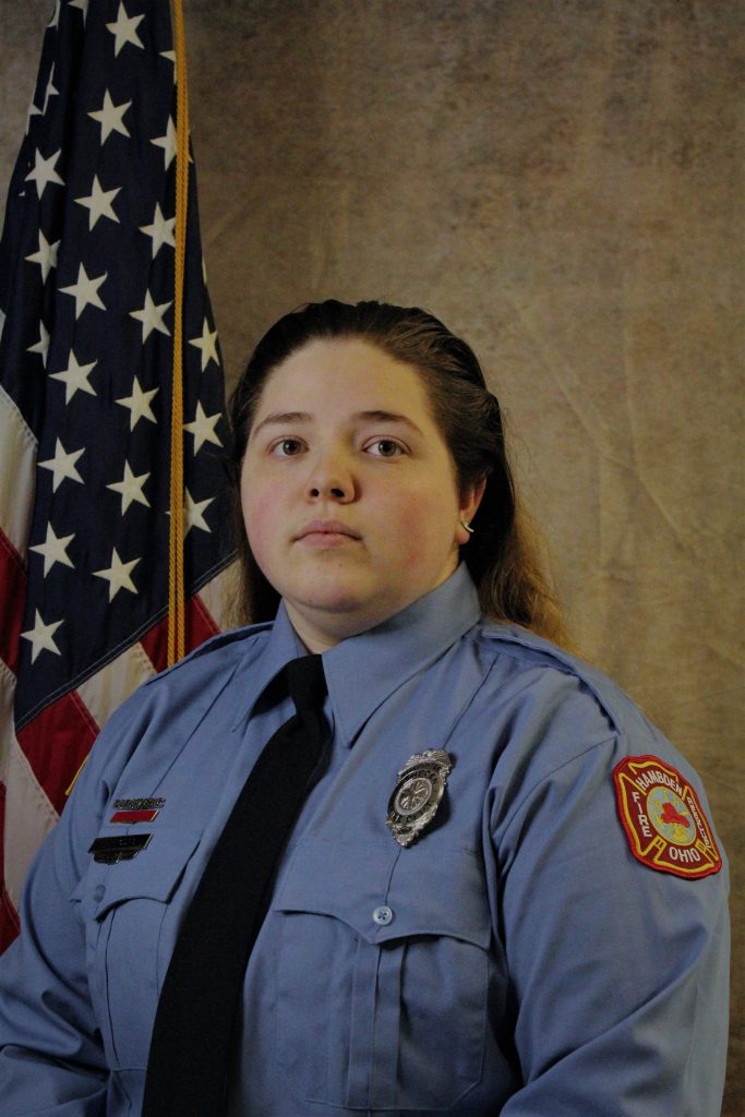 A person in a blue uniform with patches and a badge stands in front of an American flag. They have long hair and a serious expression.