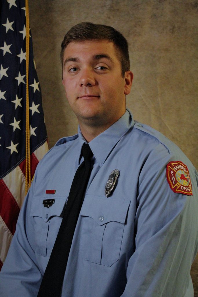 A firefighter wearing a light blue uniform with a badge and a patch featuring "Hubbard, Ohio." He stands in front of a U.S. flag, looking forward. The background is a neutral tone.