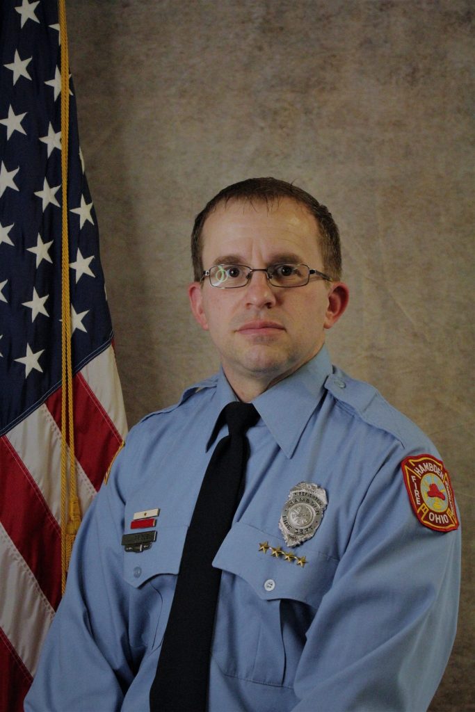 A person in a light blue uniform with badges and a tie sits in front of an American flag backdrop. The uniform has a patch on the sleeve that reads "Mansfield, Ohio.