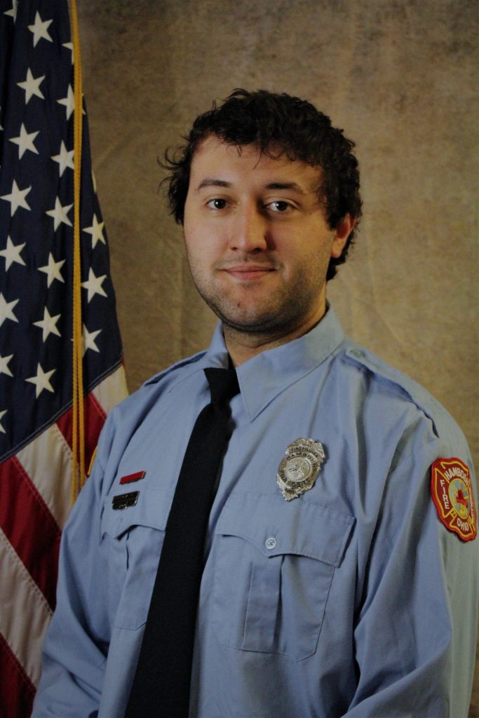 A person in a blue uniform with a fire department patch stands beside an American flag. They have a badge on their shirt and a black tie, posing against a neutral backdrop.