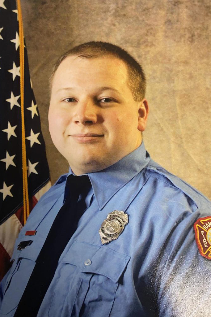 A person in a blue uniform with a badge and an American flag patch stands in front of an American flag. The background is a neutral color.