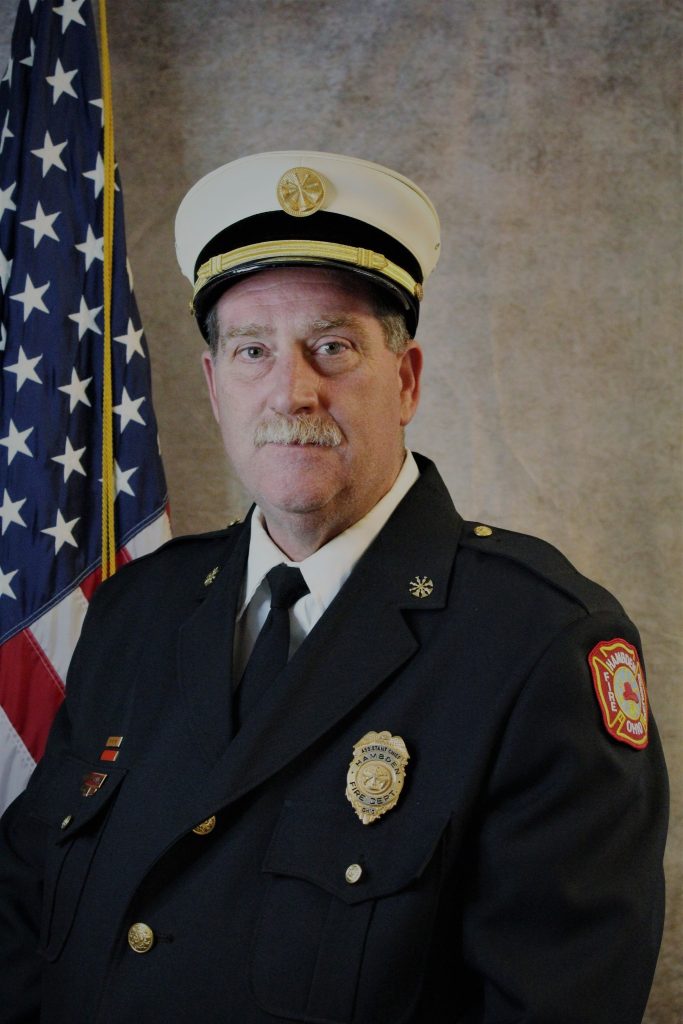 A uniformed firefighter stands next to an American flag. The uniform includes a navy jacket with a badge and shoulder patches, and a white visor cap with an emblem on the front. The background is neutral.