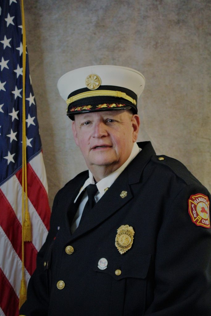 A person in a firefighter's dress uniform with badges and a white cap stands in front of an American flag. The individual has a solemn expression and the uniform bears fire department insignia.