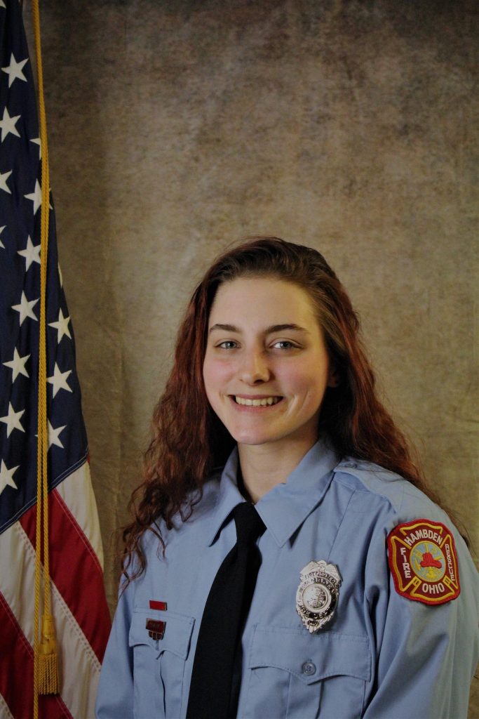 A person in a light blue uniform with a badge and a shoulder patch that says "Ohio". They are smiling, standing in front of a United States flag and a neutral backdrop.