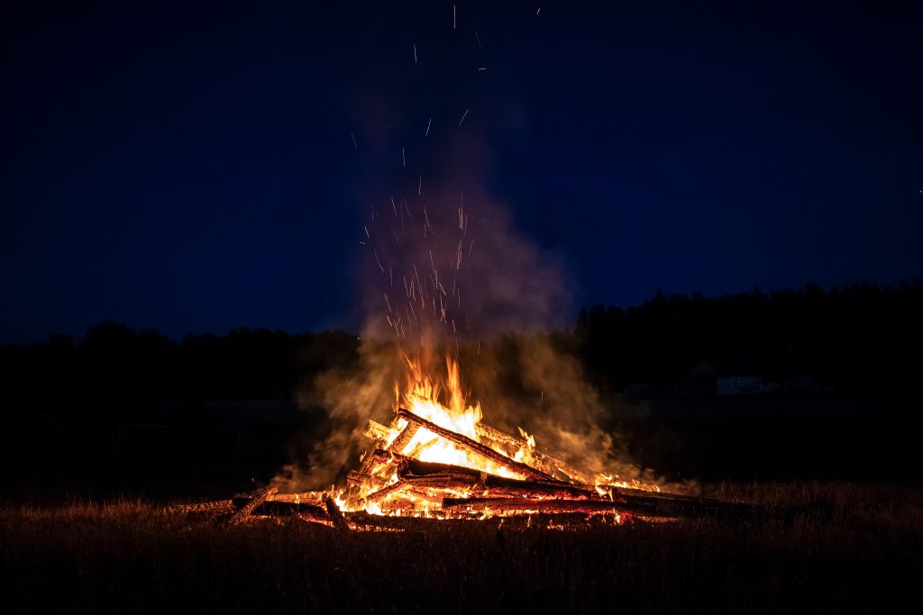 A large bonfire burns brightly against a dark blue night sky. Flames rise high, with sparks flying upwards. The surrounding area is dark, with silhouettes of trees visible in the background.