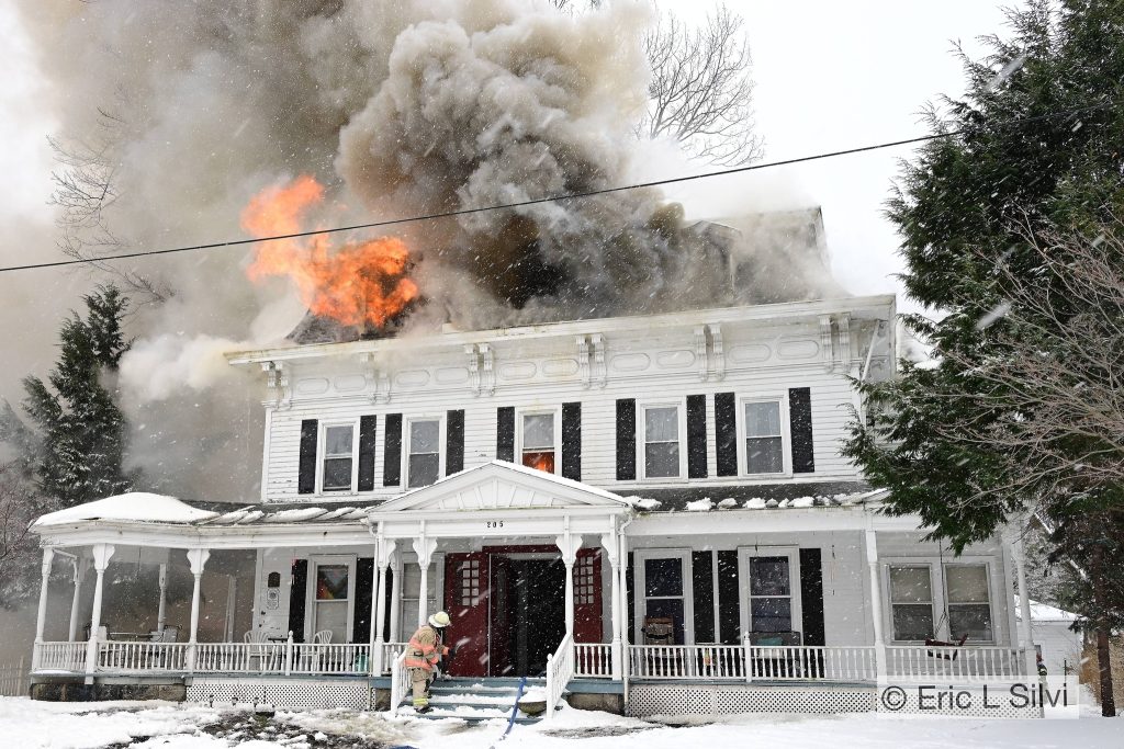 A firefighter in uniform sprays water at a large white house engulfed in flames and thick smoke. Snow covers the ground and trees are nearby.
