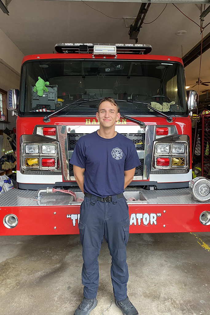 A person in a navy blue uniform stands smiling in front of a red fire truck inside a garage. There is a green stuffed toy on the front of the truck, and firefighting equipment is visible in the background.