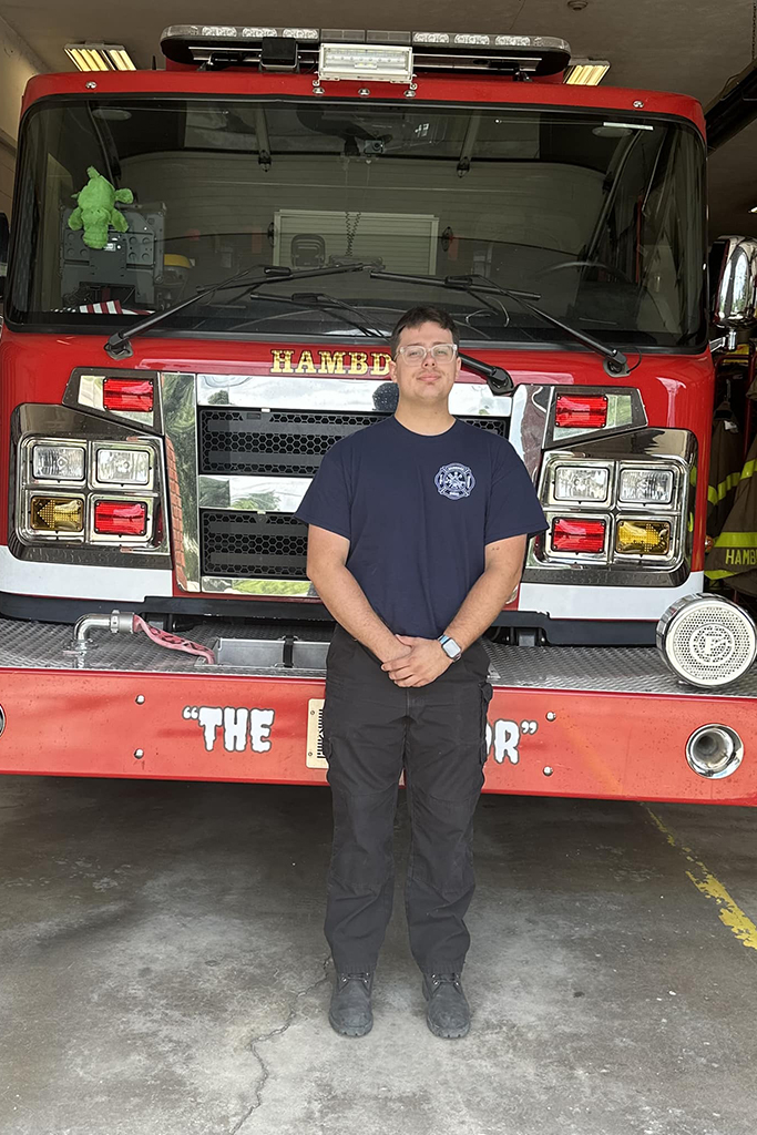 A person in a dark blue t-shirt and black pants stands in front of a red fire truck inside a garage. The truck has clear reflective features and the person has their hands clasped in front.