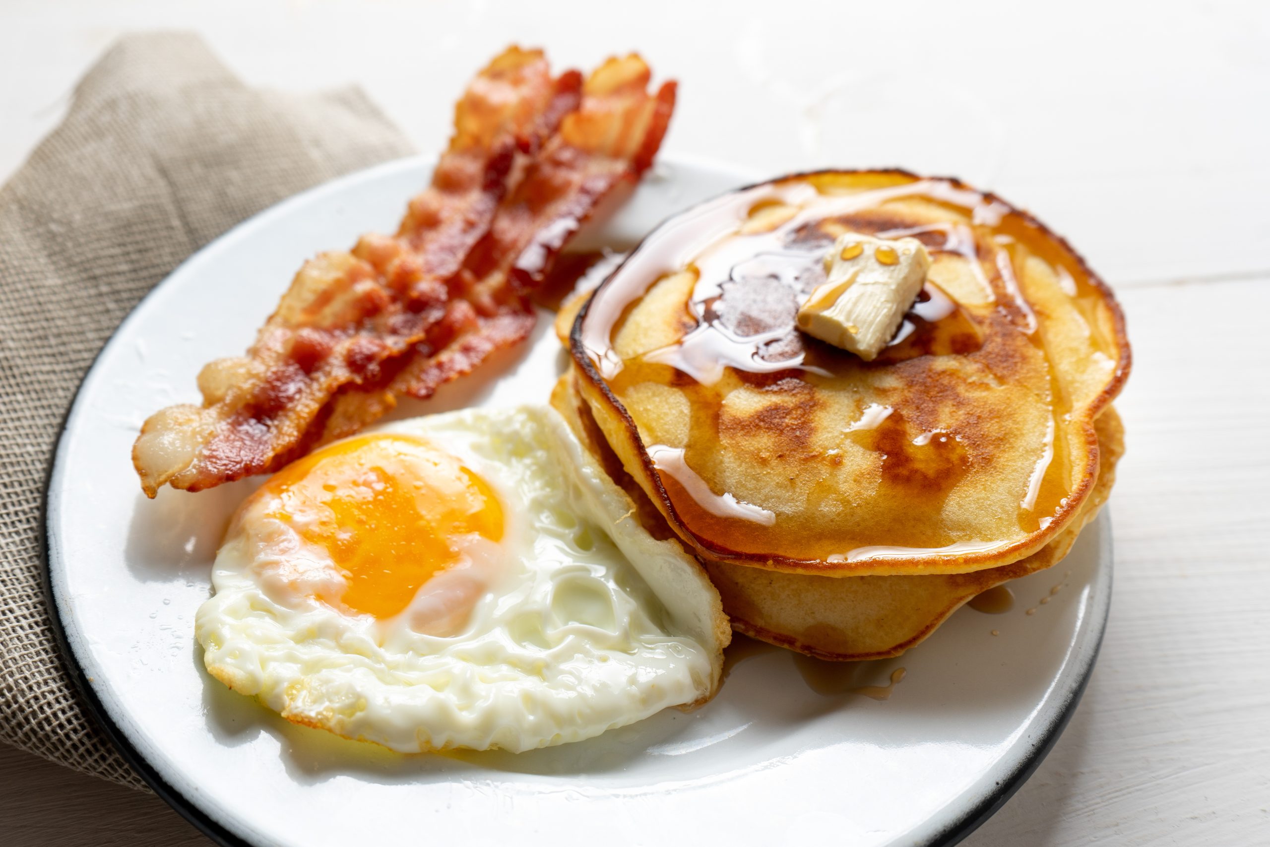 A plate with two pancakes topped with butter and syrup, two strips of crispy bacon, and a sunny-side-up egg. A beige napkin is partially visible to the side.