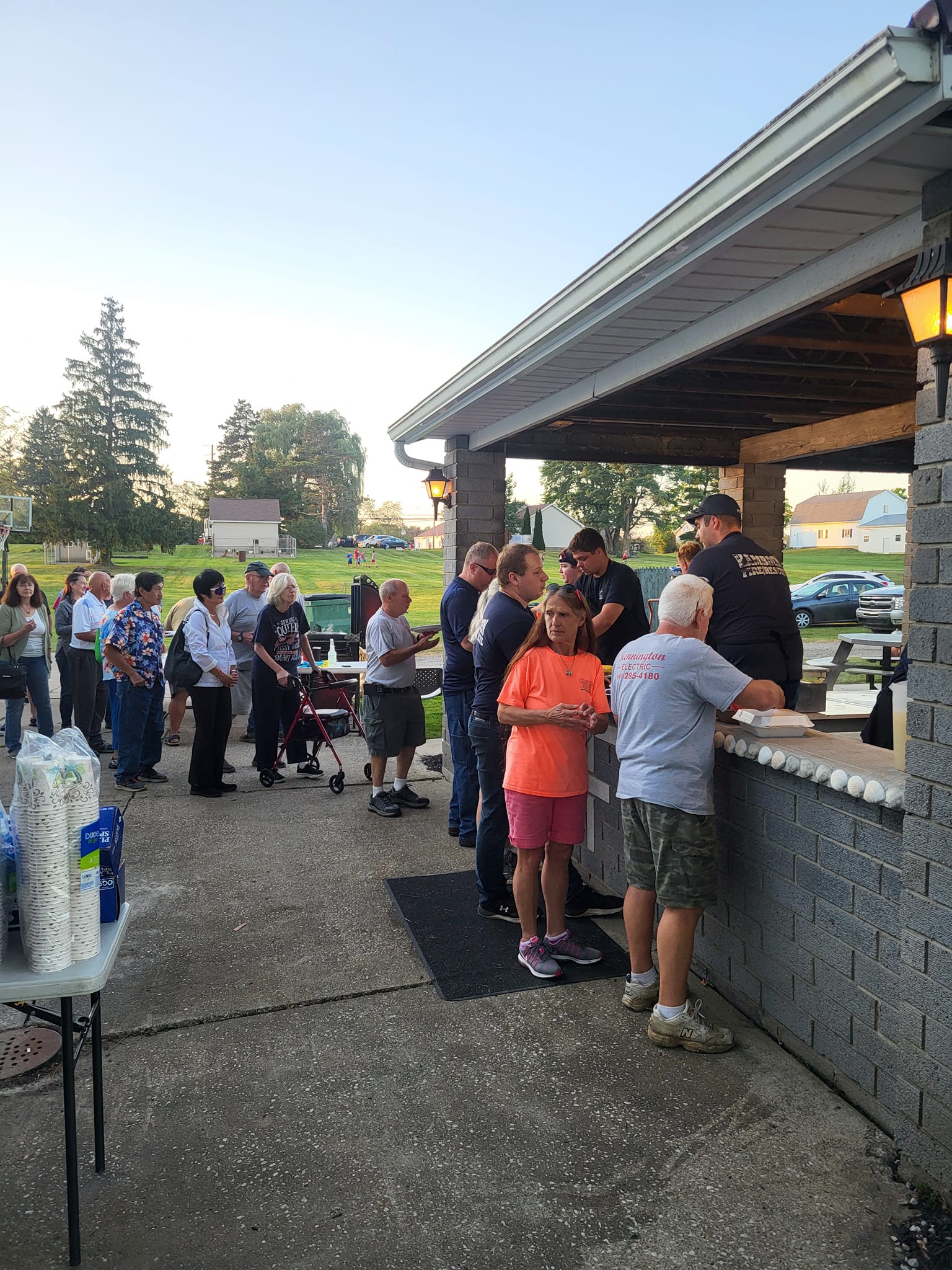 People are lining up outside a pavilion to receive food. The setting is a park with trees and houses visible in the background. Some people are holding food containers, and others stand waiting in line under a clear sky.