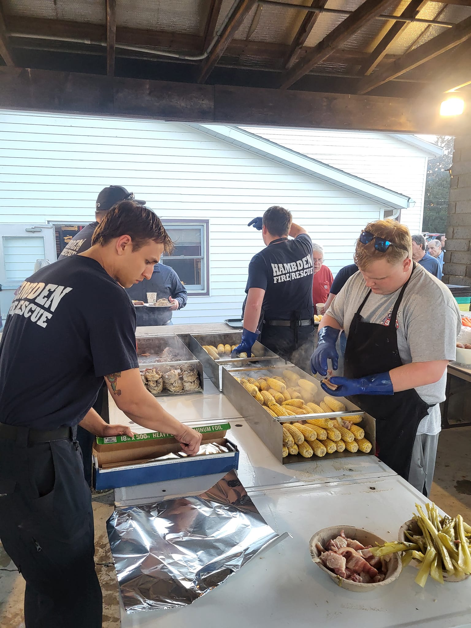 A group of people wearing "HAMDEN FIRE RESCUE" shirts prepare food. Two men are filling large trays with corn and foil-wrapped items. A tray of green beans is visible in the foreground. The setting appears to be an outdoor covered area.