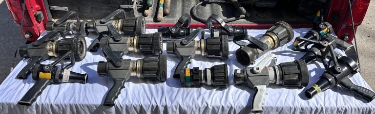 Several black and silver fire hose nozzles are arranged on a white cloth, set on the tailgate of a red vehicle with other equipment visible in the background.