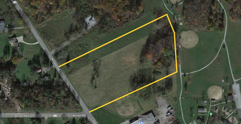 A satellite view of a grassy field outlined in yellow, bordered by Kent Road and a wooded area, with baseball fields and buildings nearby.