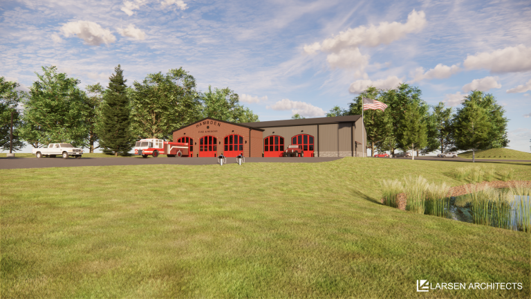 A modern fire station with red doors and fire trucks parked outside sits among green trees under a blue sky. An American flag waves nearby, and a pond is visible in the foreground.